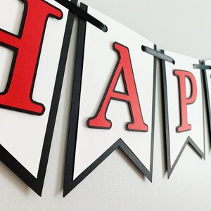 Op de afbeelding: Een rood-zwarte banner met het woord "HAPPY" in grote, vetgedrukte, rode letters. De banner is gemaakt van papier en heeft een witte achtergrond.