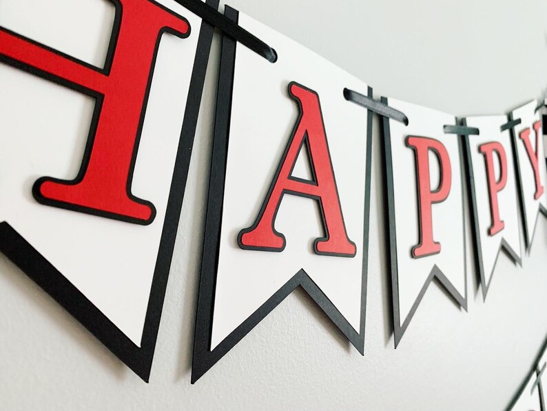Op de afbeelding: Een rood-zwarte banner met het woord "HAPPY" in grote, vetgedrukte, rode letters. De banner is gemaakt van papier en heeft een witte achtergrond.