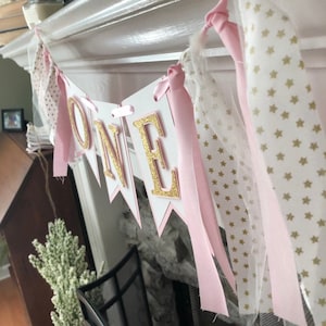 May include: A pink and gold "ONE" banner with white tulle and pink ribbon. The banner is decorated with gold stars.