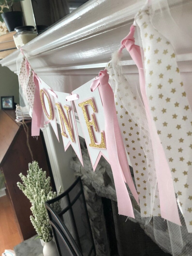 May include: A pink and gold "ONE" banner with white tulle and pink ribbon. The banner is decorated with gold stars.