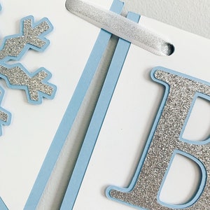 May include: A blue and white banner with a silver glitter snowflake and the letter "B" in silver glitter. The banner is strung together with white ribbon.