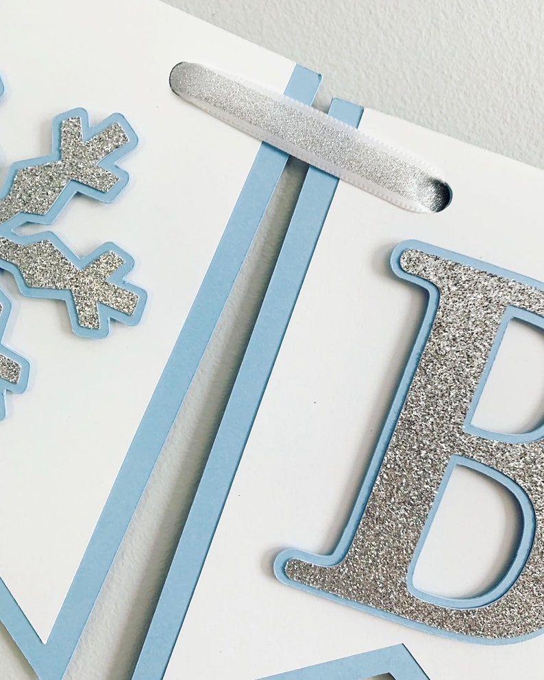 May include: A blue and white banner with a silver glitter snowflake and the letter "B" in silver glitter. The banner is strung together with white ribbon.