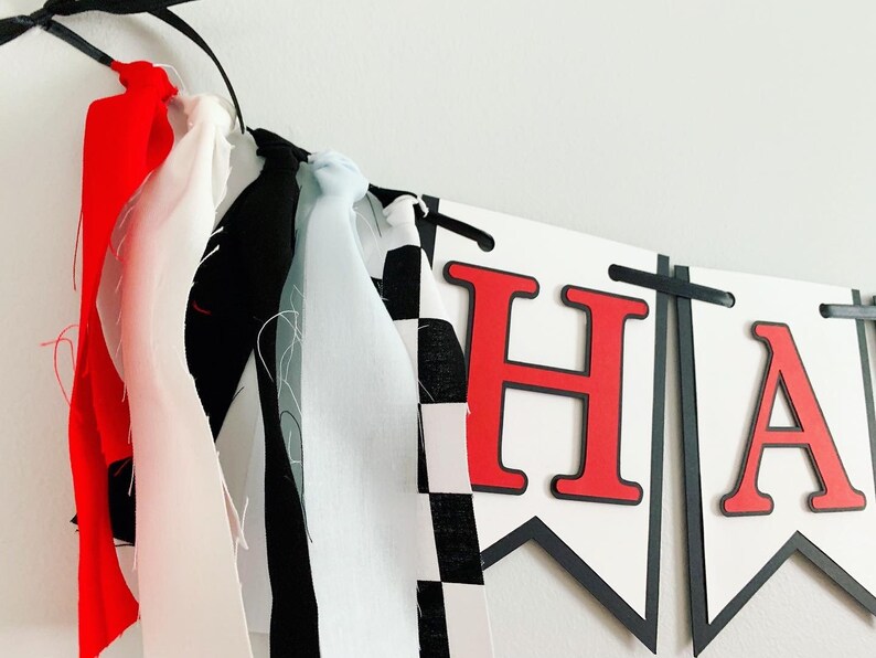 Op de afbeelding: Een witte banner met zwarte bies en rode letters die "Happy" spellen. De banner is versierd met zwart-wit geruit stof, rode, witte en blauwe stoffen streamers.