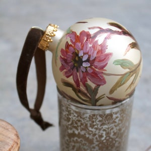 May include: A cream-colored Christmas ornament with a hand-painted pink and purple flower design. The ornament has a gold cap and a brown velvet ribbon. It rests on a decorative glass votive holder.