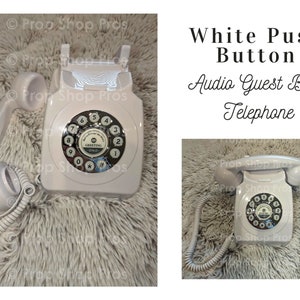 May include: White rotary dial phone with a push button for recording audio messages. The phone has a white cord and is on a white fuzzy surface. The text "Greeting" is visible on the phone's dial.