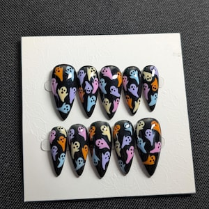 May include: A set of press-on nails with a black base and a pattern of colorful cartoon ghosts. The ghosts are in shades of orange, purple, pink, and blue. The nails are almond-shaped and arranged on a white card.