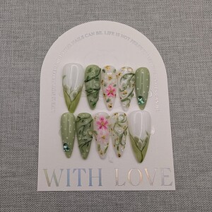 May include: A set of ten artificial nails in various shades of green and white, decorated with floral designs, leaves, and rhinestones. The nails are arranged on a white card with the words "WITH LOVE" and "LIFE IS NOT PERFECT BUT YOUR NAILS CAN BE."