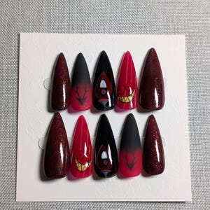 May include: A set of ten long, pointed false nails. The nails feature a variety of designs, including glittery burgundy, black with red ombre, and red with cartoon character faces. The nails are arranged on a white card.