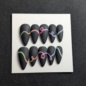 May include: A set of ten matte black press-on nails. Each nail features a colorful, curved design in shades of pink, yellow, blue, and green. The nails are arranged on a white card, perfect for showcasing nail art.