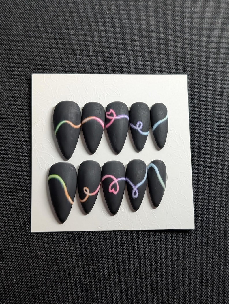 May include: A set of ten matte black press-on nails. Each nail features a colorful, curved design in shades of pink, yellow, blue, and green. The nails are arranged on a white card, perfect for showcasing nail art.