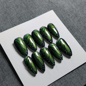 May include: Set of ten green and black press-on nails with a butterfly wing design. The nails are oval-shaped with a glossy finish. The butterfly wing design features black veins and white dots.