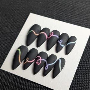May include: A set of ten matte black, almond-shaped press-on nails. Each nail features a pastel-colored, swirled design. The nails are arranged on a white, textured card.