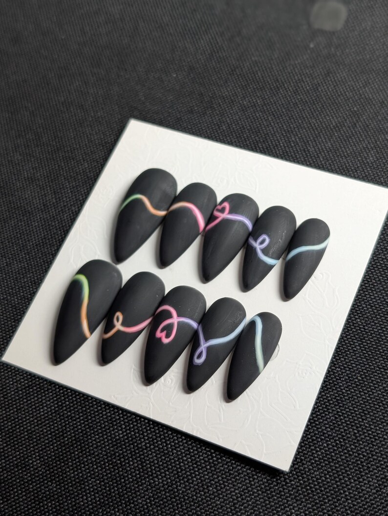 May include: A set of ten matte black, almond-shaped press-on nails. Each nail features a pastel-colored, swirled design. The nails are arranged on a white, textured card.