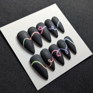 May include: A set of matte black, almond-shaped press-on nails. Each nail features a curved, colorful design in shades of green, orange, pink, and blue. The nails are arranged on a white card.