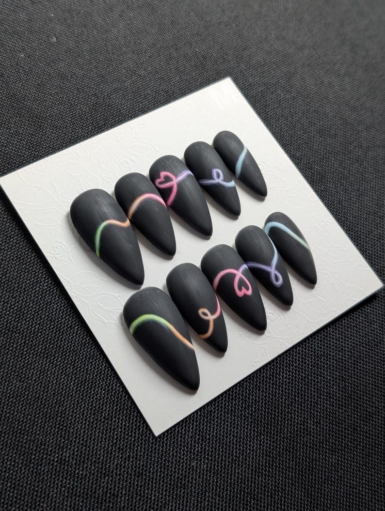 May include: A set of matte black, almond-shaped press-on nails. Each nail features a curved, colorful design in shades of green, orange, pink, and blue. The nails are arranged on a white card.