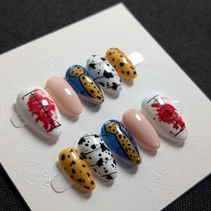 May include: A set of ten artificial nails with various designs. The designs include a red abstract pattern on white, a solid nude color, a blue and gold pattern, a black and white spotted pattern, and a yellow and black spotted pattern.