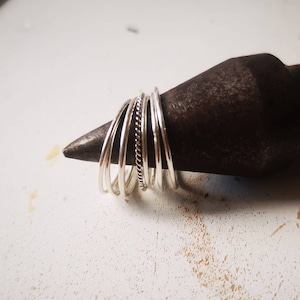 May include: A stack of silver rings, some plain and some with a textured design, are displayed on a dark metal surface.