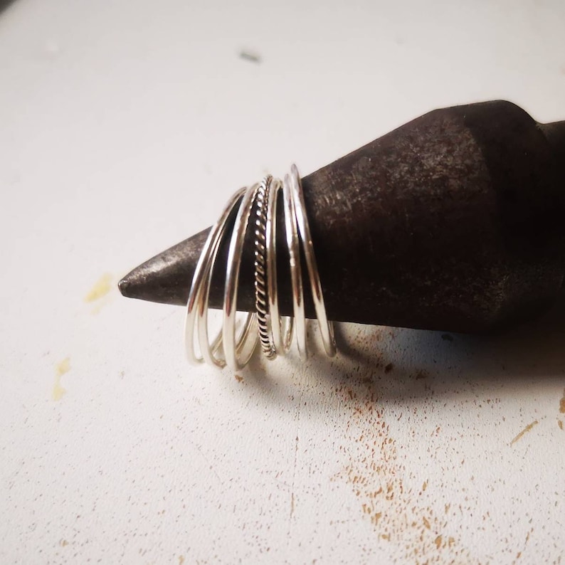 May include: A stack of silver rings, some plain and some with a textured design, are displayed on a dark metal surface.