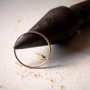 May include: A silver hoop earring is resting on a black metal tool. The tool is shaped like a cone and has a rough texture.