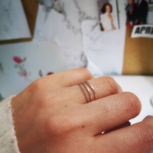 May include: Two silver rings on a finger. The ring on the bottom is a simple band, while the ring on top is a twisted band.