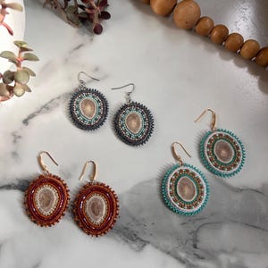 May include: Three pairs of circular dangle earrings featuring central polished antler slices surrounded by intricate beadwork. One pair has dark grey beads, one has rust-coloured beads, and one has bright turquoise and white beads with gold-toned hooks.