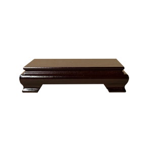 Puede incluir: Un pedestal de madera marrón oscuro con una parte superior rectangular y patas curvas. El pedestal está hecho de madera pulida y tiene un acabado liso.