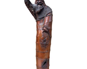May include: A wooden sculpture of a smiling, bearded man with an outstretched arm. The figure is carved from a tree trunk, with detailed carvings of fish and clothing. The sculpture is primarily brown with darker accents.