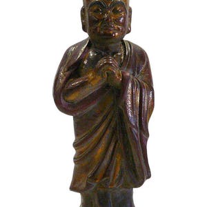 May include: A small, dark brown wooden statue of a monk with a gold-painted face. The monk is wearing a robe and has his hands clasped in prayer.