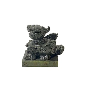Puede incluir: Una escultura de jade negro de un león guardián mítico chino, también conocido como foo dog, sentado sobre una base de jade verde con detalles dorados.
