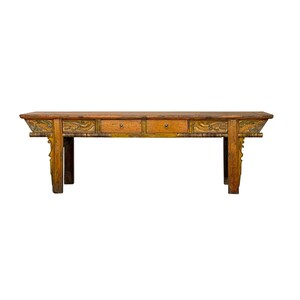 May include: A long, rectangular wooden console table with a weathered mustard-yellow finish. The front features two central drawers with small circular metal pulls, flanked by decorative carved panels depicting dragon motifs on each side.
