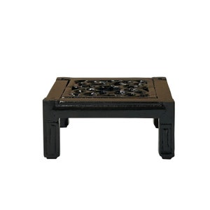Puede incluir: Una pequeña mesa cuadrada de madera negra con detalles florales tallados en la parte superior. La mesa tiene cuatro patas cortas y rectangulares y está sobre un fondo blanco. La mesa mide aproximadamente 25 cm de ancho.