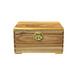 May include: A small, wooden box with a natural wood grain finish. The box features a brass-colored clasp and hinges. The corners are dovetailed for added strength. The box is rectangular and sits on small feet.