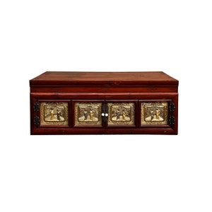 May include: A dark red wooden chest with four doors. Each door has a brass panel with a raised design of figures in a landscape. The chest has a flat top and is decorated with a simple design.