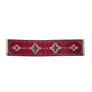 May include: A long, rectangular red runner rug with a detailed floral pattern. The rug features a central design with repeating cross-like motifs in white, navy, and light blue. The edges are finished with a fringe.