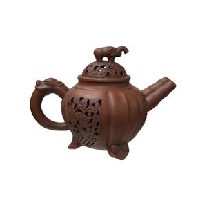May include: Brown ceramic teapot with intricate floral cutouts and a dragon-shaped handle. The lid features a small elephant figurine. The spout has a bamboo-like design. The teapot is designed with three feet.