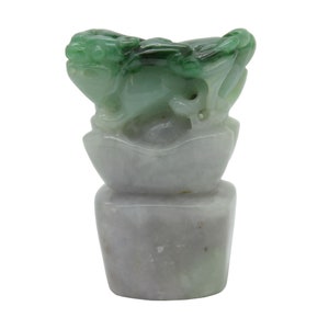 Può includere: Una scultura in giada di una creatura mitologica, forse un drago o un leone, seduta su un fiore di loto. La scultura è verde e bianca, con dettagli intricati.