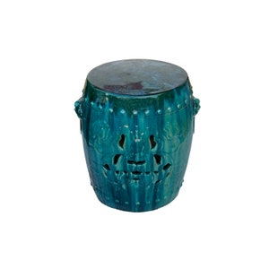 May include: A teal ceramic garden stool with a round top and a drum-like shape. The stool features decorative cut-out patterns and is adorned with small, raised studs around the top and bottom. It has two ornate handles on the sides.