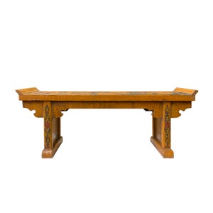 Puede incluir: Un banco de madera con un acabado marrón claro y diseños florales pintados intrincados en tonos de rojo, azul y verde. El banco tiene una parte superior rectangular y dos patas a cada lado, con un borde superior ligeramente curvado.