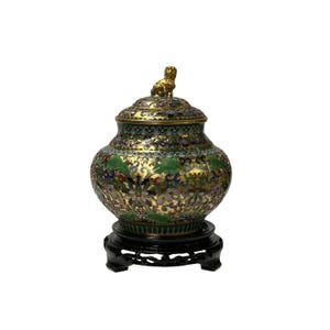 May include: An ornate, antique cloisonné jar with a lid, featuring a gold-colored lion finial. The jar is decorated with green, blue, and purple floral patterns on a gold background. It sits on a black wooden stand.