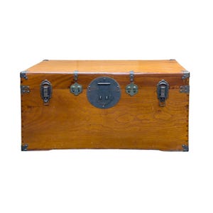 May include: A rectangular wooden storage chest with a natural light brown wood grain finish. The chest features dark metal corner reinforcements, two side latches, and a central circular metal locking plate with a horizontal pull handle.