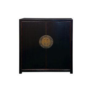 May include: A rectangular black cabinet with two doors featuring a circular brass plate in the center. The cabinet has a smooth, dark finish with subtle red trim along the edges and a decorative, intricate metal design on the central hardware.