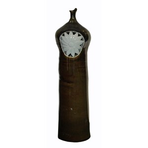 May include: A tall, brown ceramic vase with a white ceramic flower design on the front. The vase has a narrow neck and a wide base.
