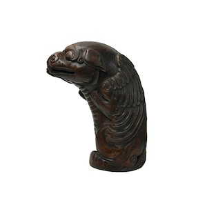 Może przedstawiać: Rzeźba z rzeźbionego drewna przedstawiająca mityczne stworzenie z psią głową, skrzydłami i zakrzywionym ciałem. Rzeźba jest w kolorze ciemnobrązowym i wydaje się być wykonana z drewna. Stworzenie jest w pozycji siedzącej.