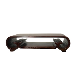May include: A dark brown, wooden coffee table with a unique, curved design. The table features a rectangular top and a lower shelf, supported by curved legs. The wood has a glossy finish, adding to its elegant appearance.