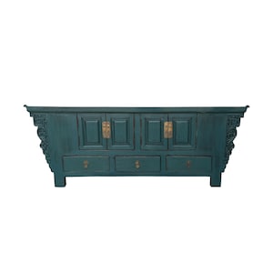 May include: A teal-colored wooden sideboard featuring four cabinet doors with brass hardware and three lower drawers with circular brass pulls. The piece has flared ends with intricate carved scrollwork details on the side supports.