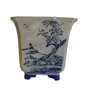 May include: A square ceramic planter with a blue and white landscape design. The planter has four blue feet and a slightly flared rim.