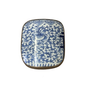 Puede incluir: Una pequeña caja de porcelana rectangular con un diseño floral azul y blanco. La caja tiene un borde dorado y está decorada con un patrón chino tradicional.