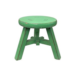 May include: A small, round, green wooden stool with three legs. The stool has a distressed, painted finish, showing signs of wear. The legs are angled outwards for stability. The seat is circular and has a few small rectangular cutouts.