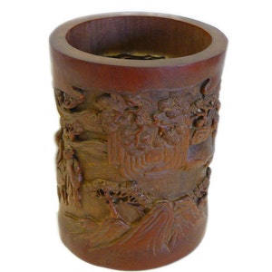 May include: A brown cylindrical bamboo vase with intricate carvings of a landscape scene. The vase is carved with trees, mountains, and figures.