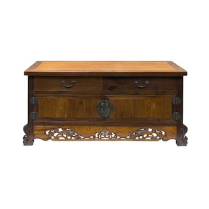 May include: A rectangular wooden cabinet featuring a light brown top surface, two upper drawers with dark metal pulls, and two lower cabinet doors with a central circular metal latch. The base includes intricate carved wooden detailing.
